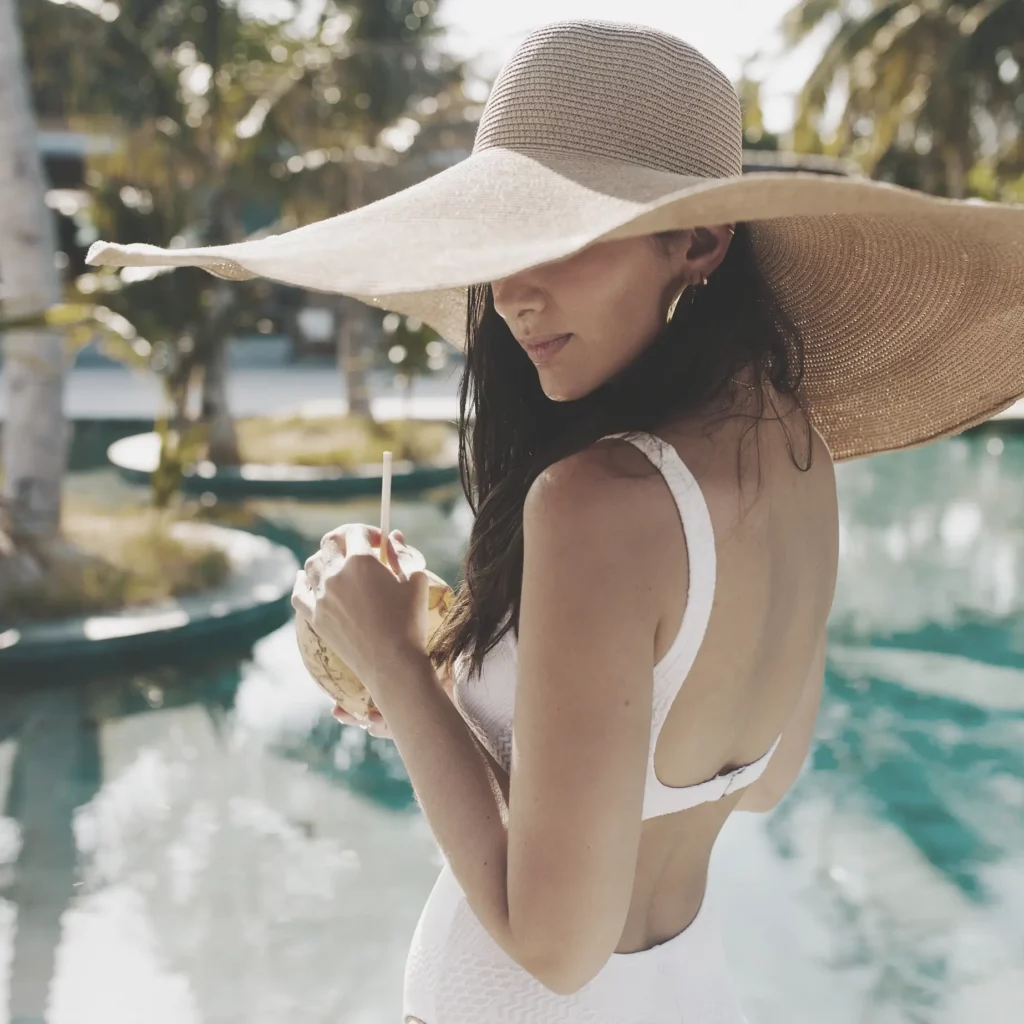 Frau im Bikini steht vor einem Pool und trinkt aus einer Kokosnuss